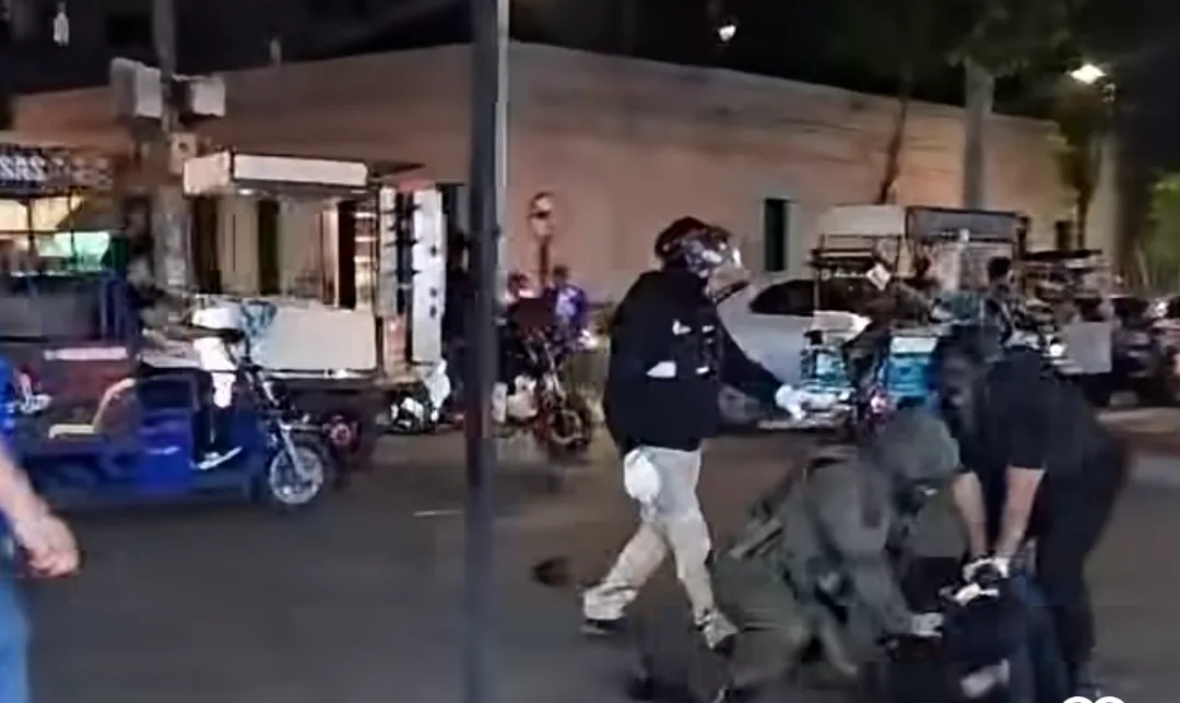 Funcionarios con casco y guantes someten a una persona en el suelo en una calle con carros de comida estacionados
