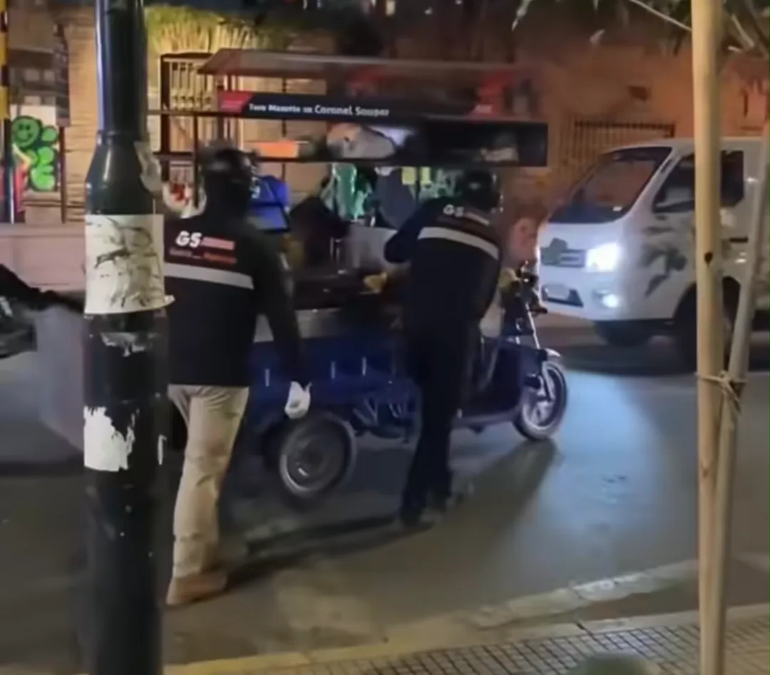 Funcionarios con chaquetas de seguridad retienen un triciclo eléctrico de trabajo en la calle de noche